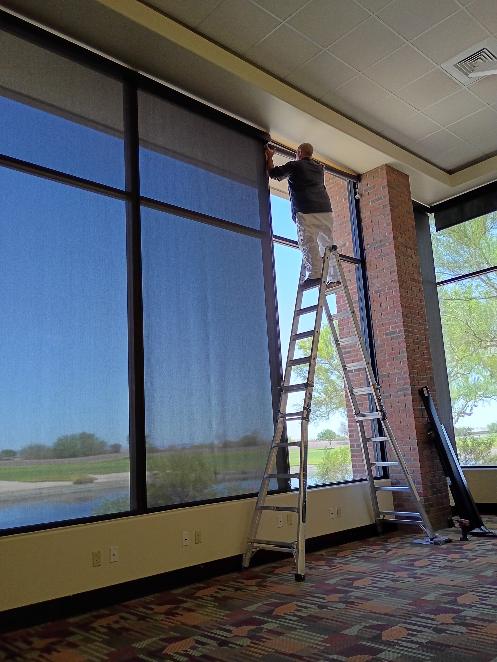 Installer on a ladder mounting a roller shade above tall commercial windows