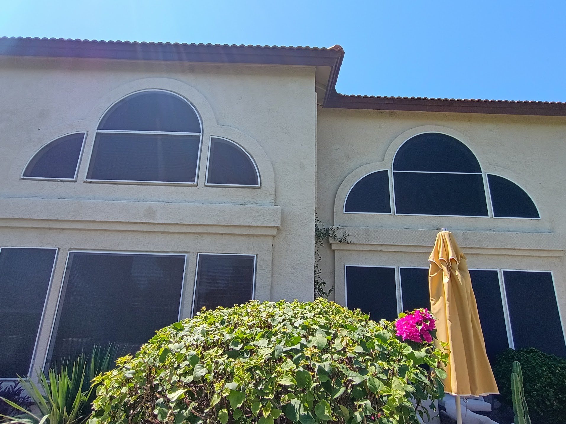 Two-story stucco home with arched upper windows, a trimmed hedge, and a yellow patio umbrella